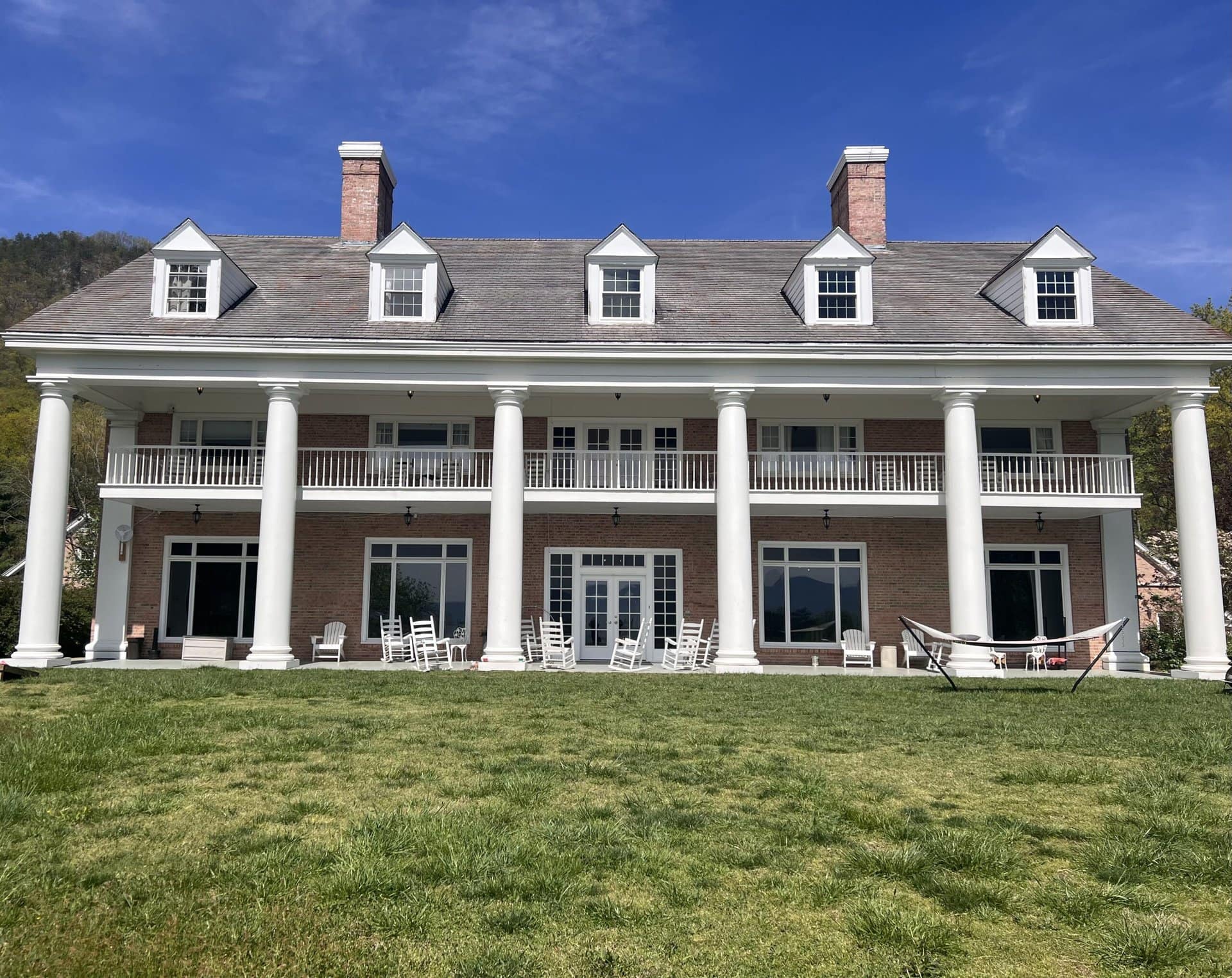 A large two-story brick mansion with white columns and a symmetrical design. It features multiple windows and chimneys, a porch with rocking chairs, and a hammock on a well-kept lawn. The sky is clear with a few wispy clouds.