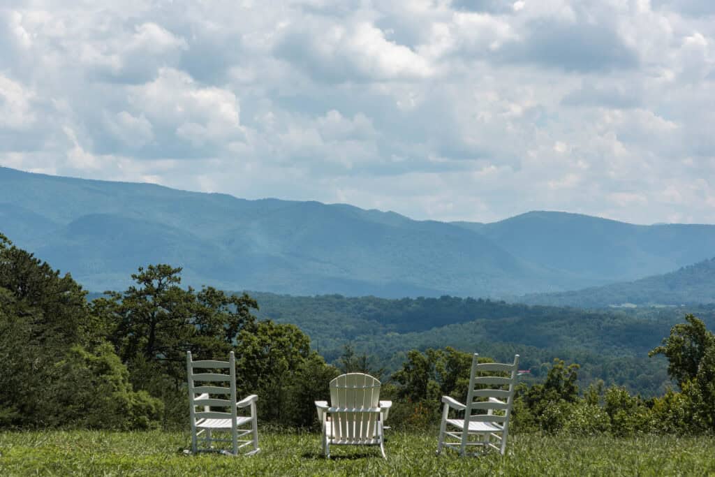 Three white chairs face lush, rolling mountains under a cloudy sky. Green trees and grass fill the foreground, providing a serene and scenic view.