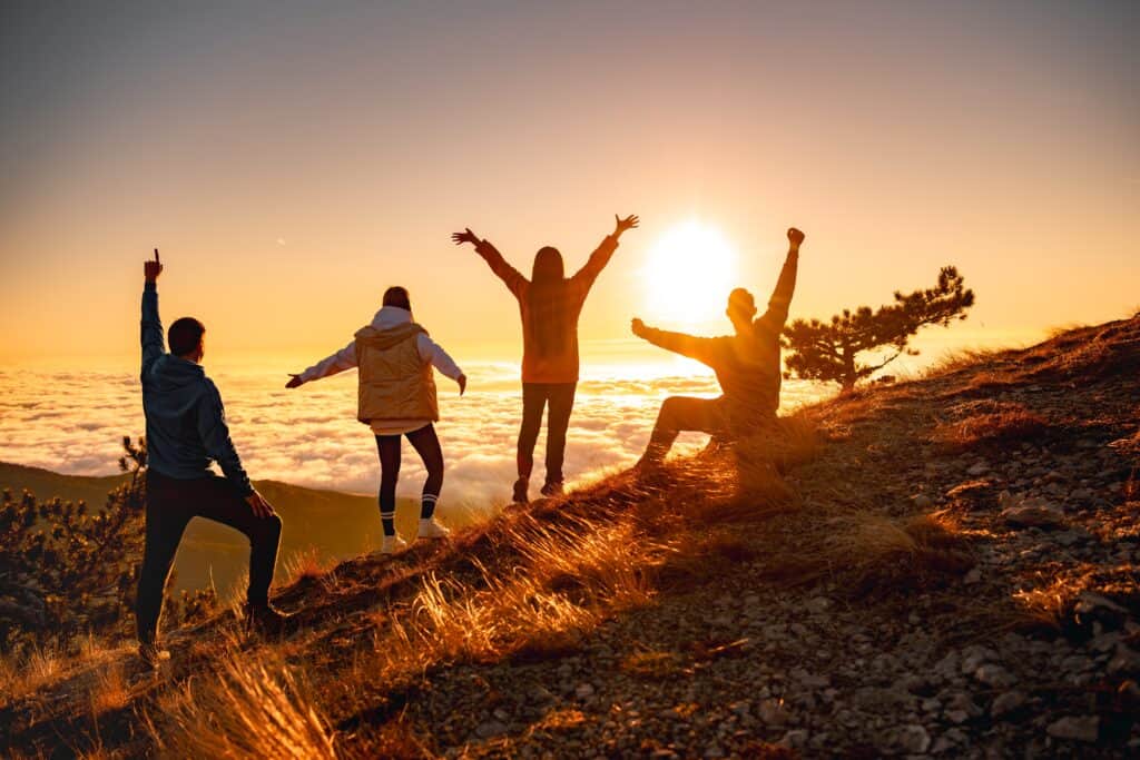 Four people stand atop a hill at sunset, with arms raised in joy. They overlook a sea of clouds below, surrounded by dry grass and sparse trees. The sky is a warm mix of orange and yellow hues.