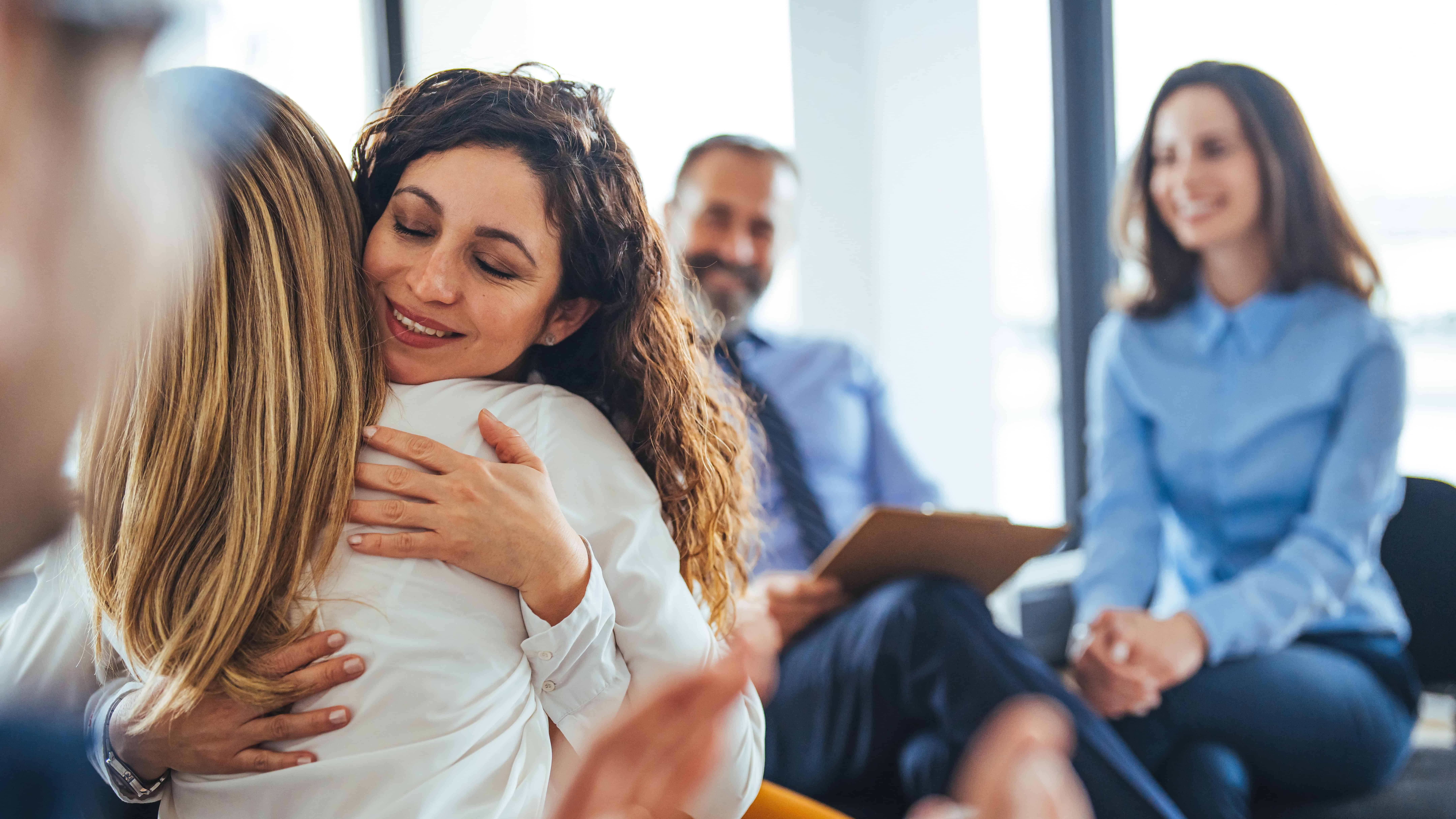 Two women are hugging in a bright office setting. Two other people, a man and a woman, are smiling and watching them in the background. The scene conveys a sense of warmth and support.