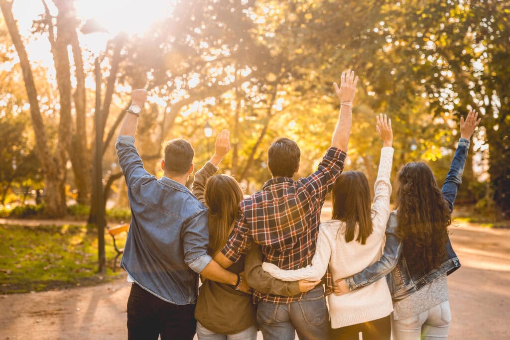 Five people with their arms around each other are raising their hands, standing on a sunlit path surrounded by trees. They are facing away, enjoying the bright, warm sunlight filtering through the leaves.