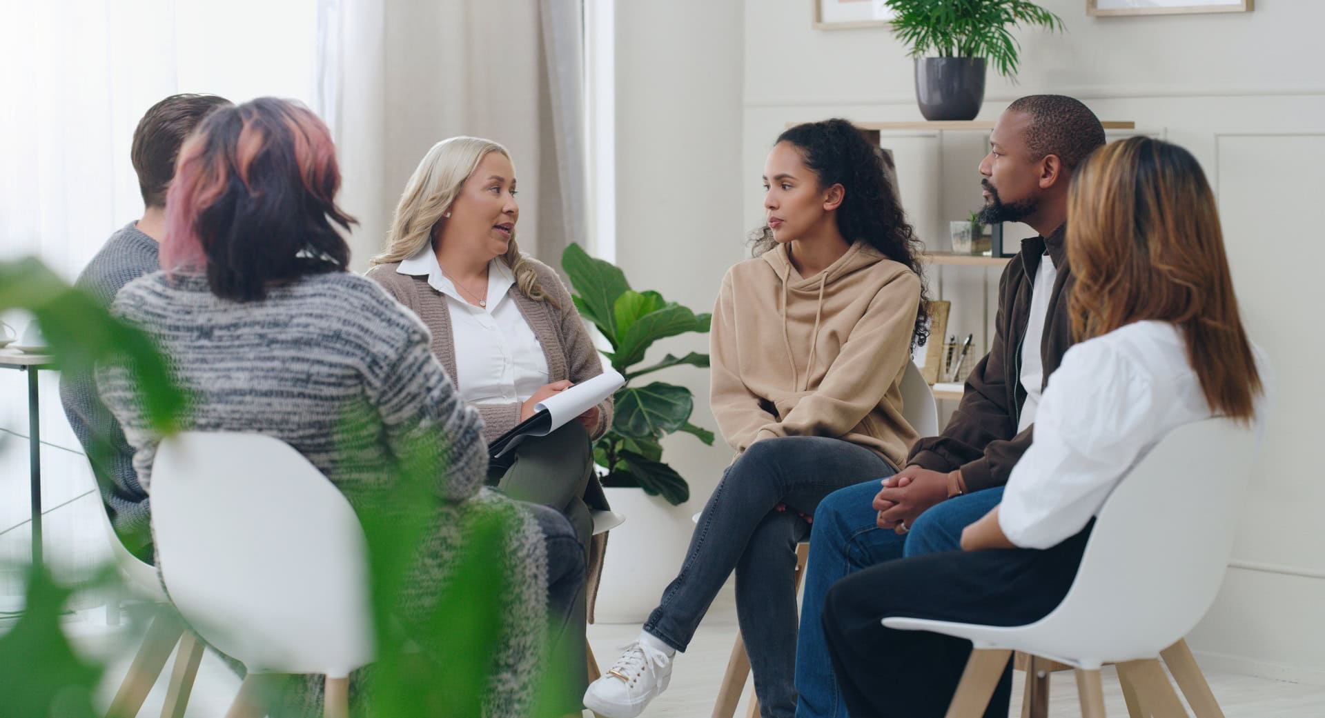 A group of five people sit in a circle in a bright room with plants. They appear to be engaged in a discussion or meeting. One woman holds a notebook, and they all face each other attentively.