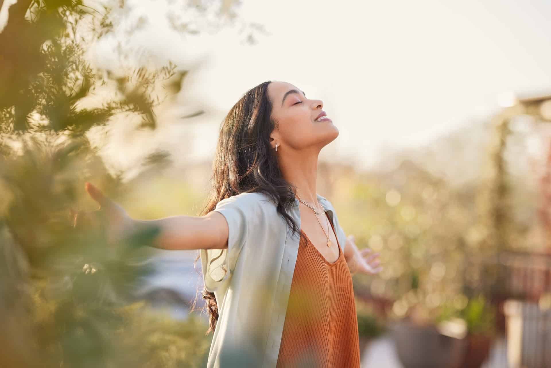 A woman with long dark hair stands outdoors with her arms outstretched and eyes closed, embracing the sunlight. She is wearing an orange top and a light shirt, surrounded by greenery with a serene expression.