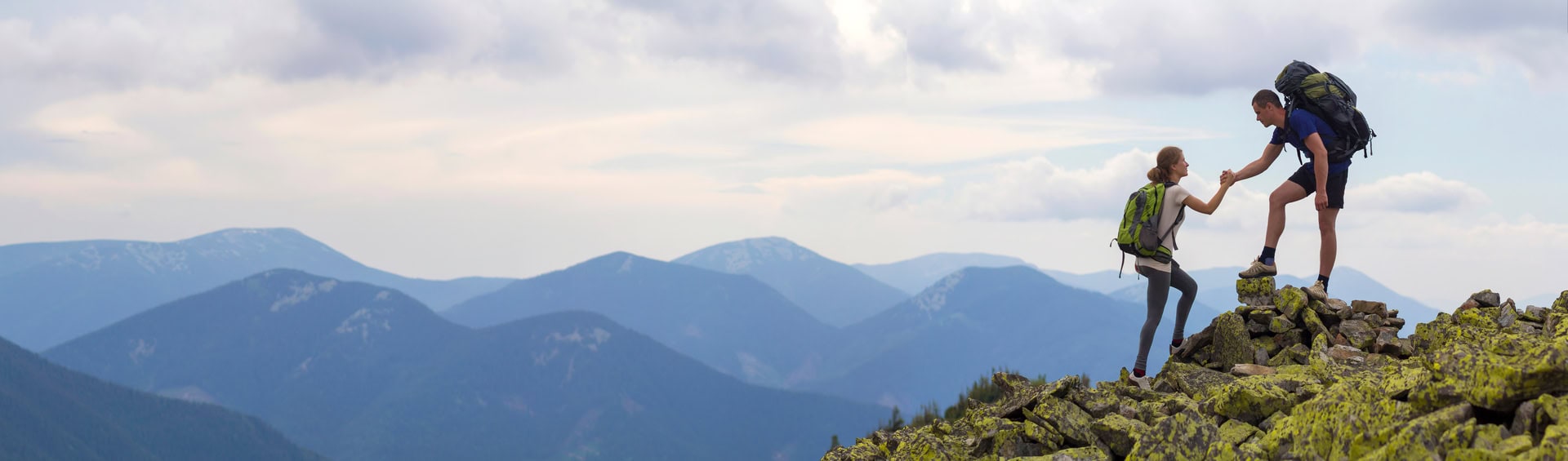 A man and woman are hiking on a rocky mountain summit. The man, standing, offers a hand to help the woman, who is kneeling. Both have backpacks. In the background, rolling mountains stretch under a partly cloudy sky.