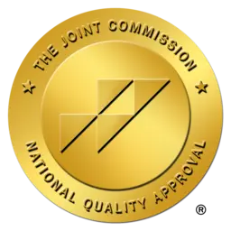 Gold circular seal with text: THE JOINT COMMISSION NATIONAL QUALITY APPROVAL. The center features an abstract logo with interlocking lines. The seal represents certification of quality standards.