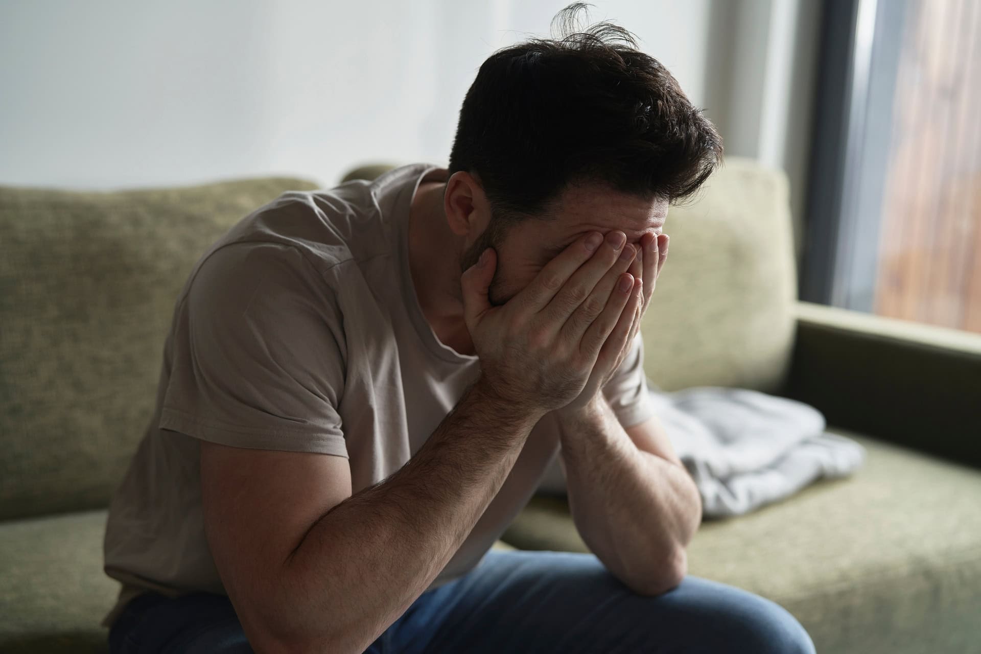 A man sits on a green sofa with his elbows on his knees, covering his face with his hands, appearing distressed or contemplative, in a softly lit room. A blanket is draped over the couch beside him.