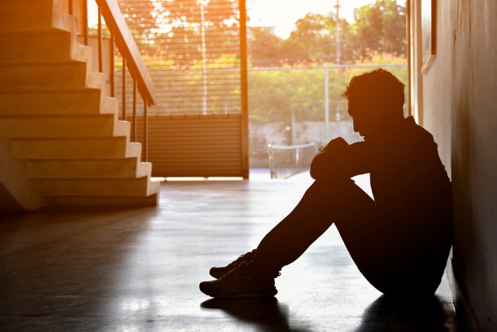 Silhouette of a person sitting on the floor against a wall, with knees drawn up and head resting on arms. Sunlight filters through a window, casting shadows on the floor. Stairs and a fenced area are visible in the background.