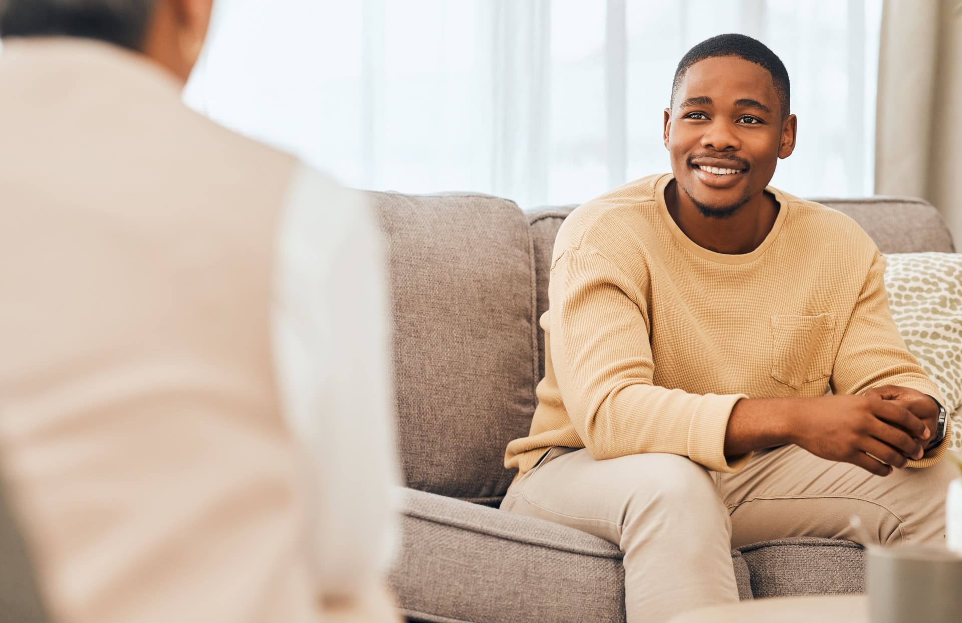 A person in a beige sweater sits on a gray couch, smiling and engaged in conversation with another person who is partially visible. The setting appears casual and comfortable.