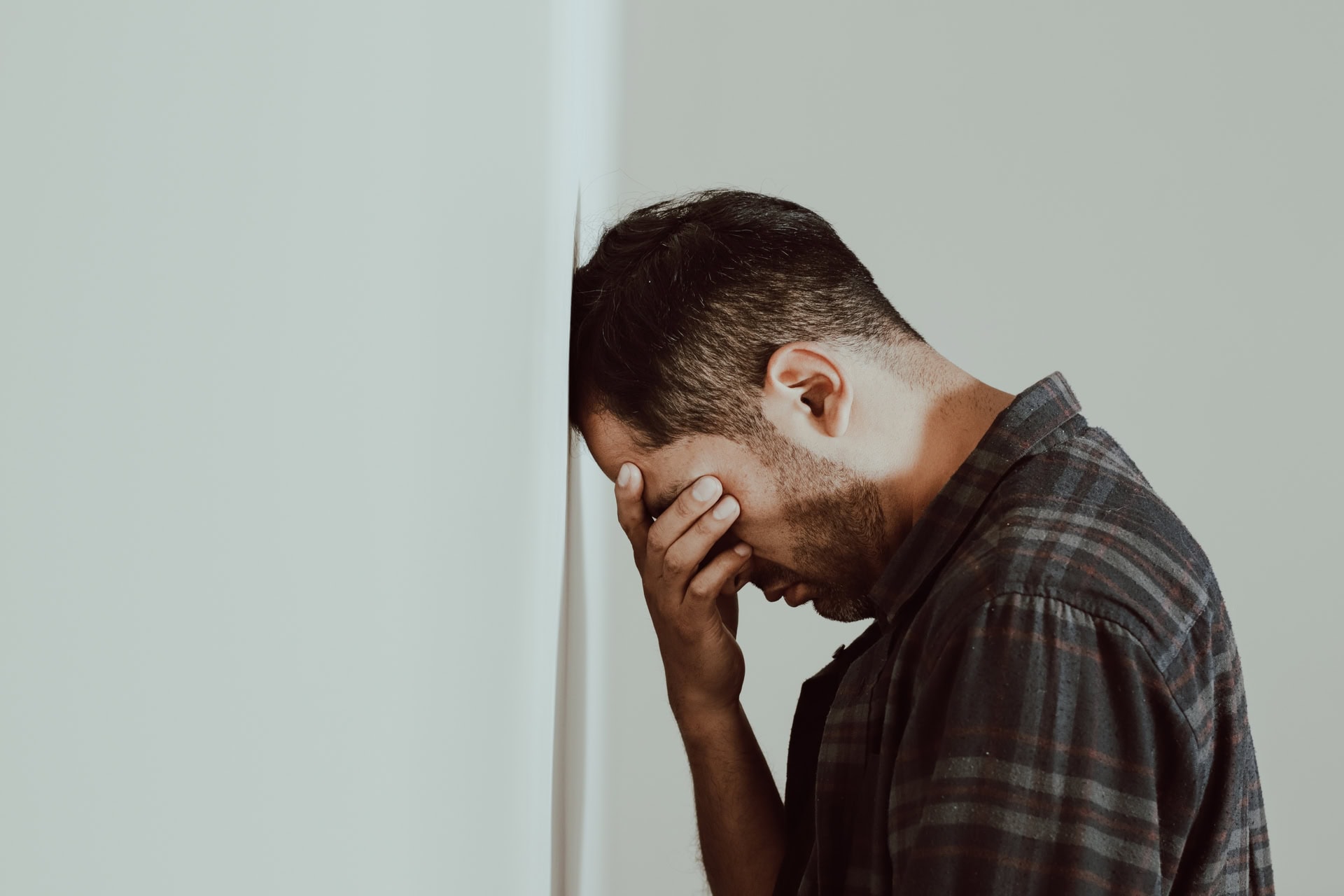 A man in a plaid shirt stands with his head resting against a white wall, covering his face with one hand. He appears to be in distress or deep thought. The atmosphere is somber and contemplative.