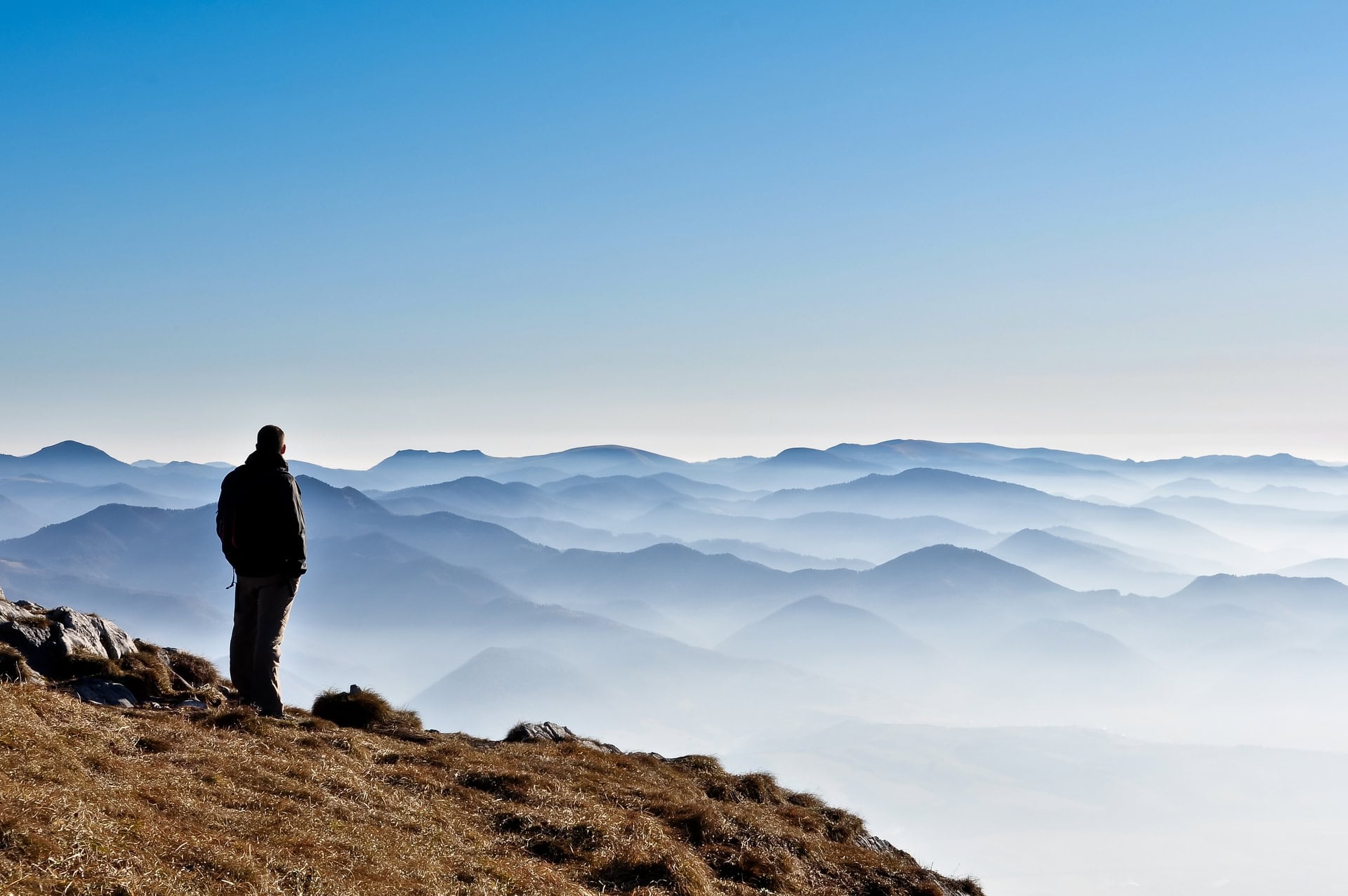 A person stands on a hilltop, gazing at a vast landscape of layered mountains under a clear blue sky. The scene conveys a sense of tranquility and wonder.