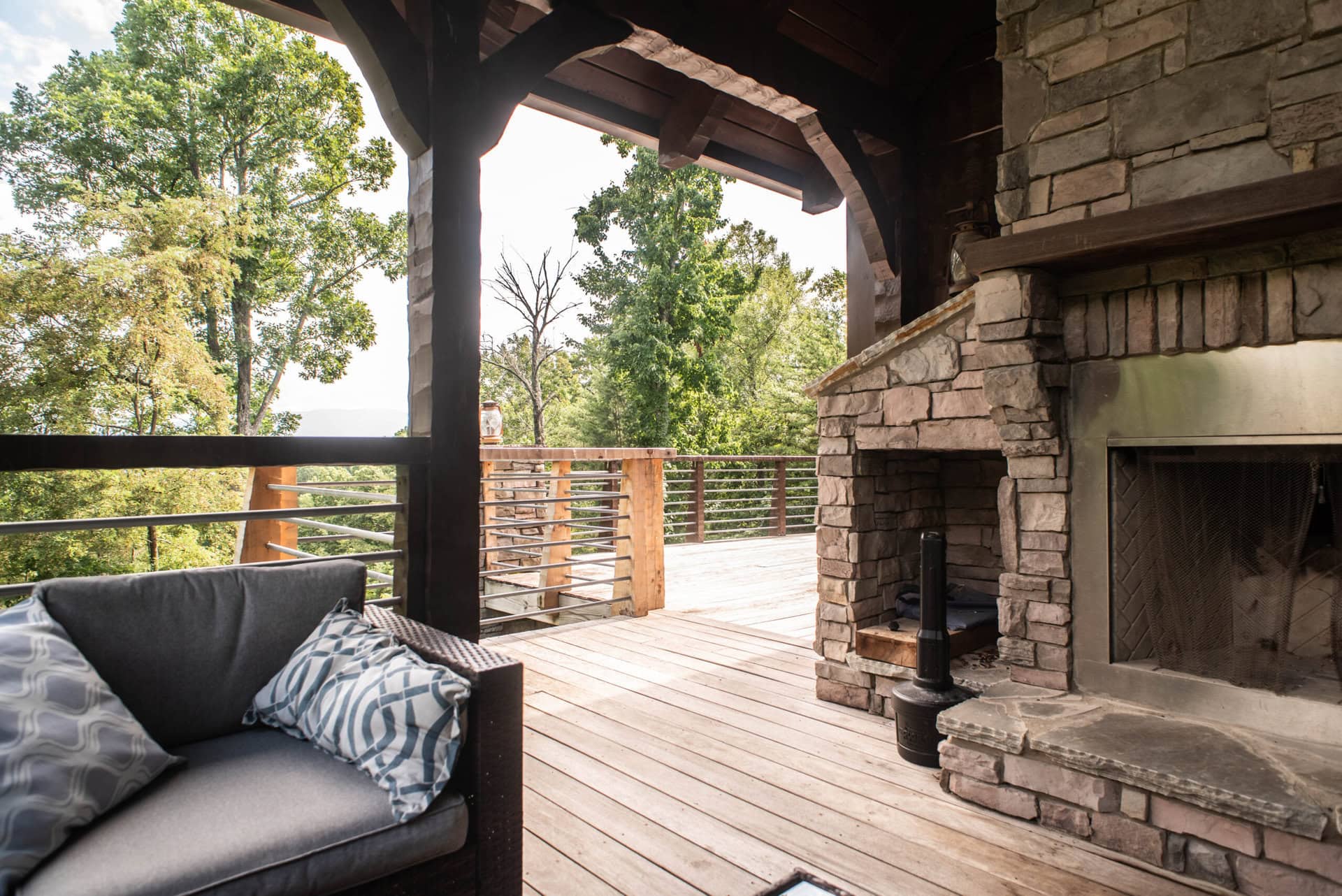 A cozy outdoor patio with a stone fireplace, wooden beams, and a comfortable gray couch with patterned pillows. The area overlooks a lush forest view on a bright day.