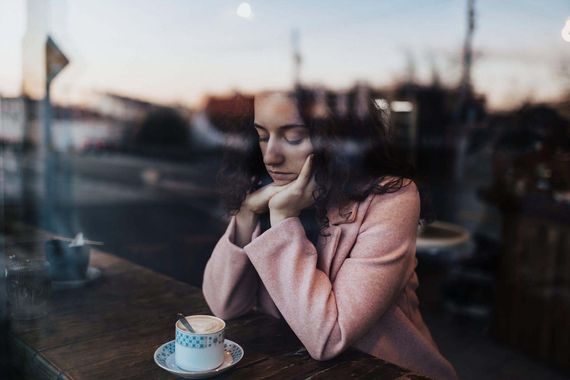 A person in a pink coat sits alone at a café table, looking down thoughtfully. A cup of coffee is in front of them. The image is taken through a window, reflecting the street scene outside. The atmosphere is calm and contemplative.