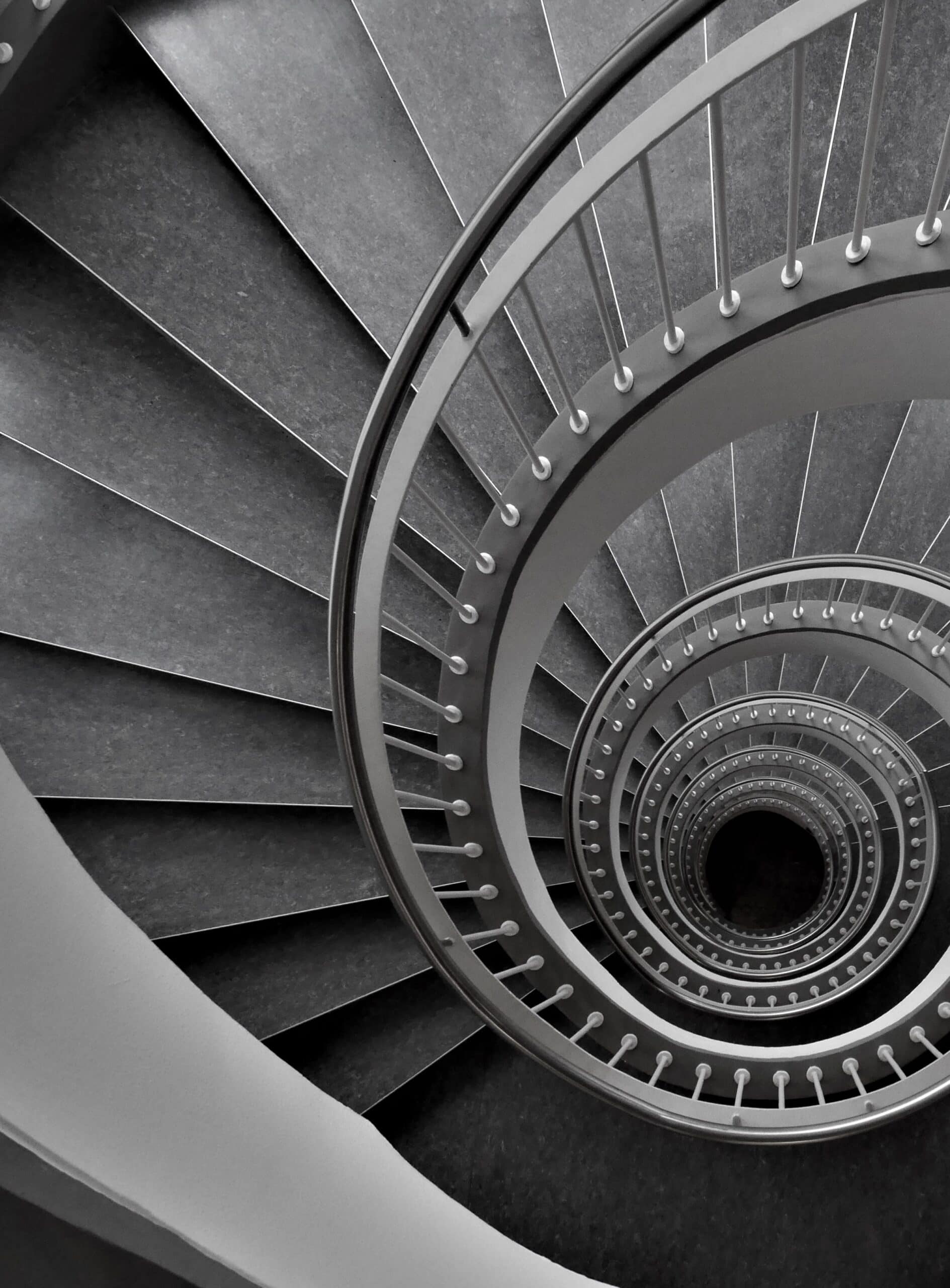 A spiral staircase made of gray metal seen from above. The steps curve downward in a circular motion, creating a hypnotic pattern that leads to a black center. The railing features vertical balusters at regular intervals.