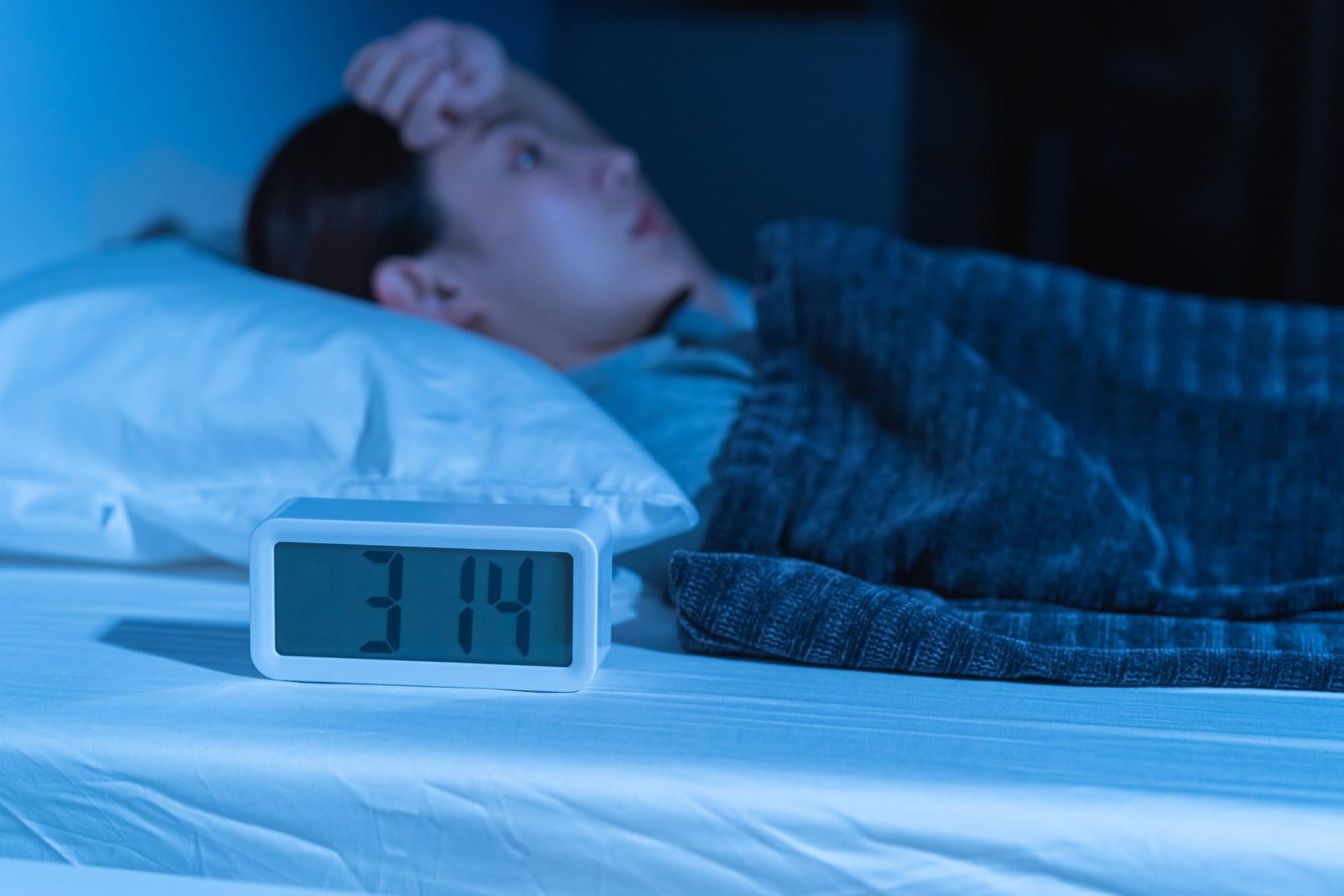 A person lies awake in bed at night, partially covered by a gray blanket, with an anxious expression. An alarm clock on the bedside table displays 3:14, casting a blue glow in the dimly lit room.