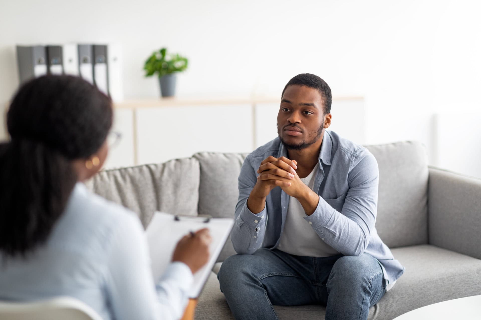 A man sits on a gray couch, attentively listening to a therapist holding a clipboard. The setting appears to be a light, modern office with a plant and binders in the background.