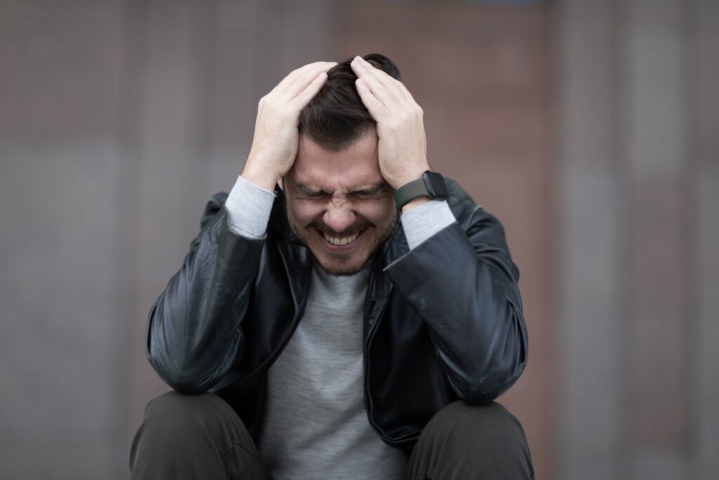 A man in a leather jacket is sitting with his hands on his head, eyes closed, and a pained expression. The background is blurred, focusing on his expression of distress or frustration.