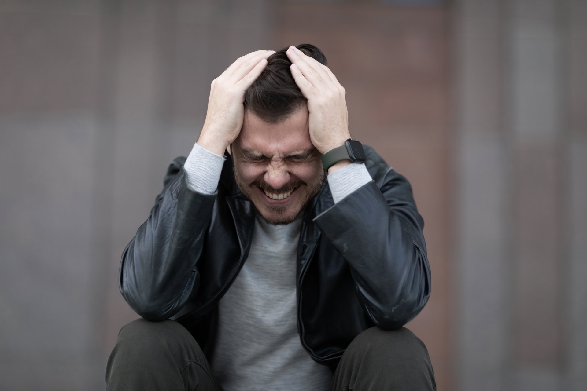 A man in a leather jacket is sitting with his hands on his head, eyes closed, and a pained expression. The background is blurred, focusing on his expression of distress or frustration.