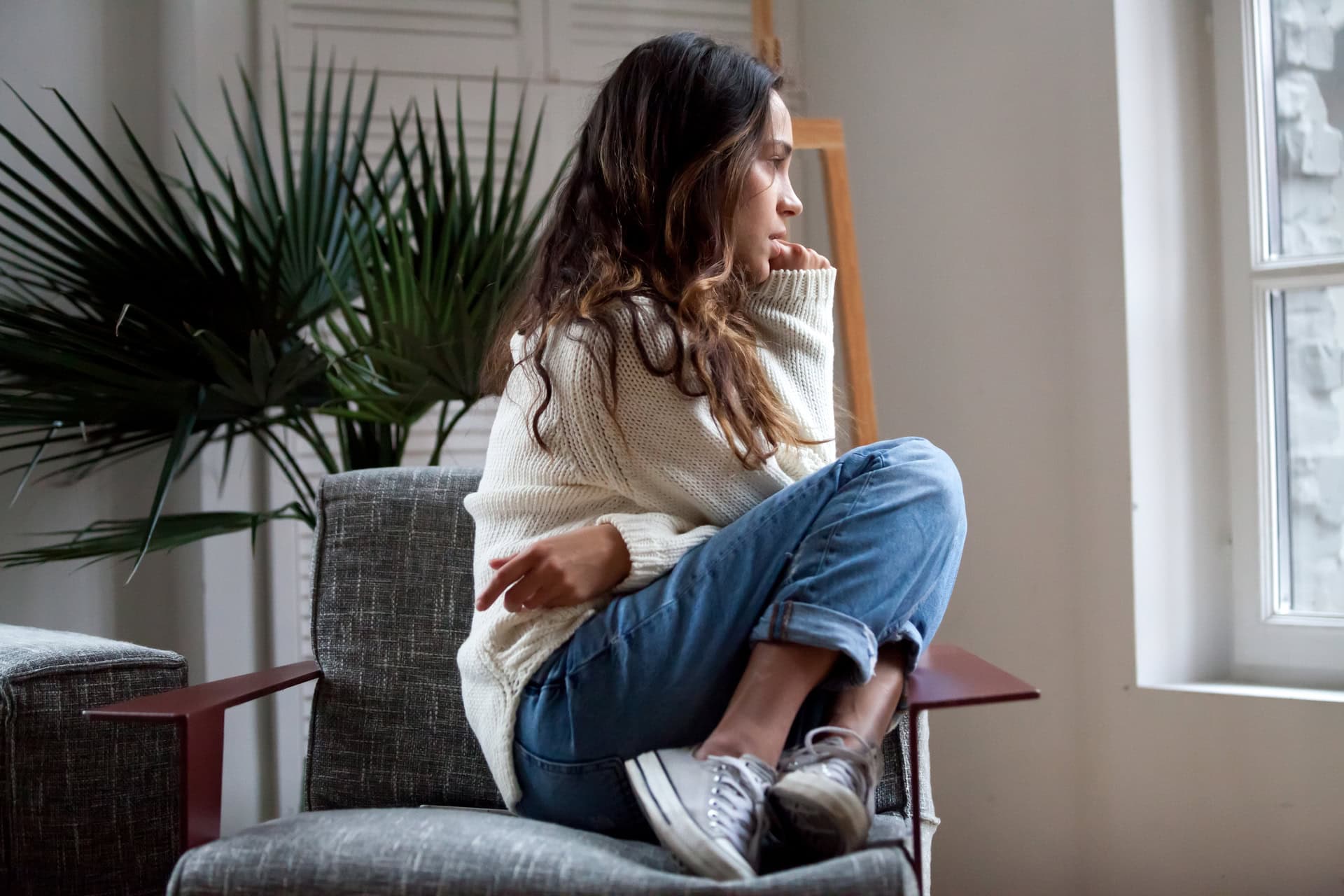 A woman with long hair, wearing a cream sweater and jeans, sits curled up on a chair, gazing thoughtfully out a window. A green plant is in the background, adding a touch of nature to the cozy indoor setting.