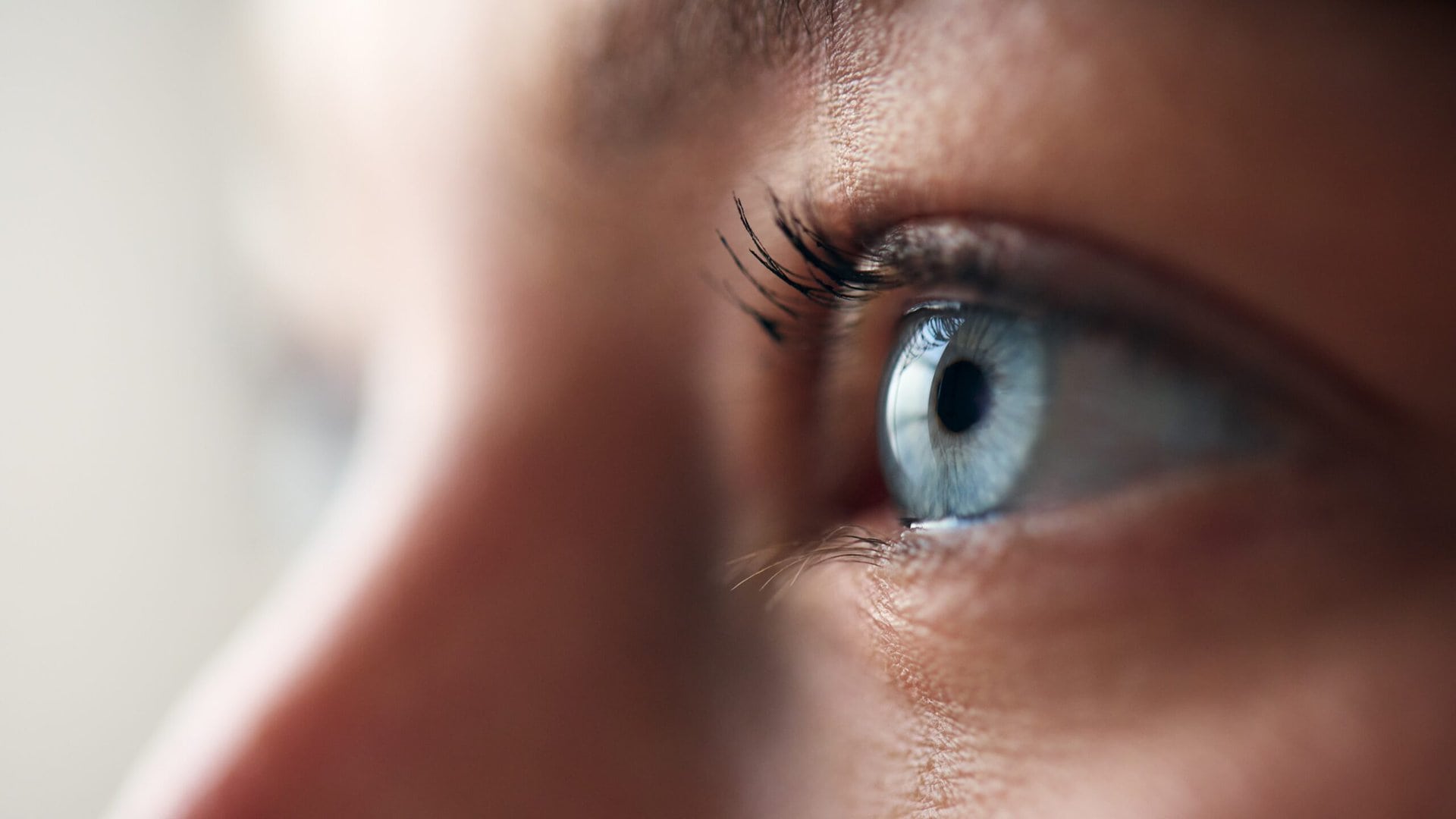 Close-up of a persons blue eye looking to the side. The focus is on the eye with visible eyelashes and a soft, blurred background.