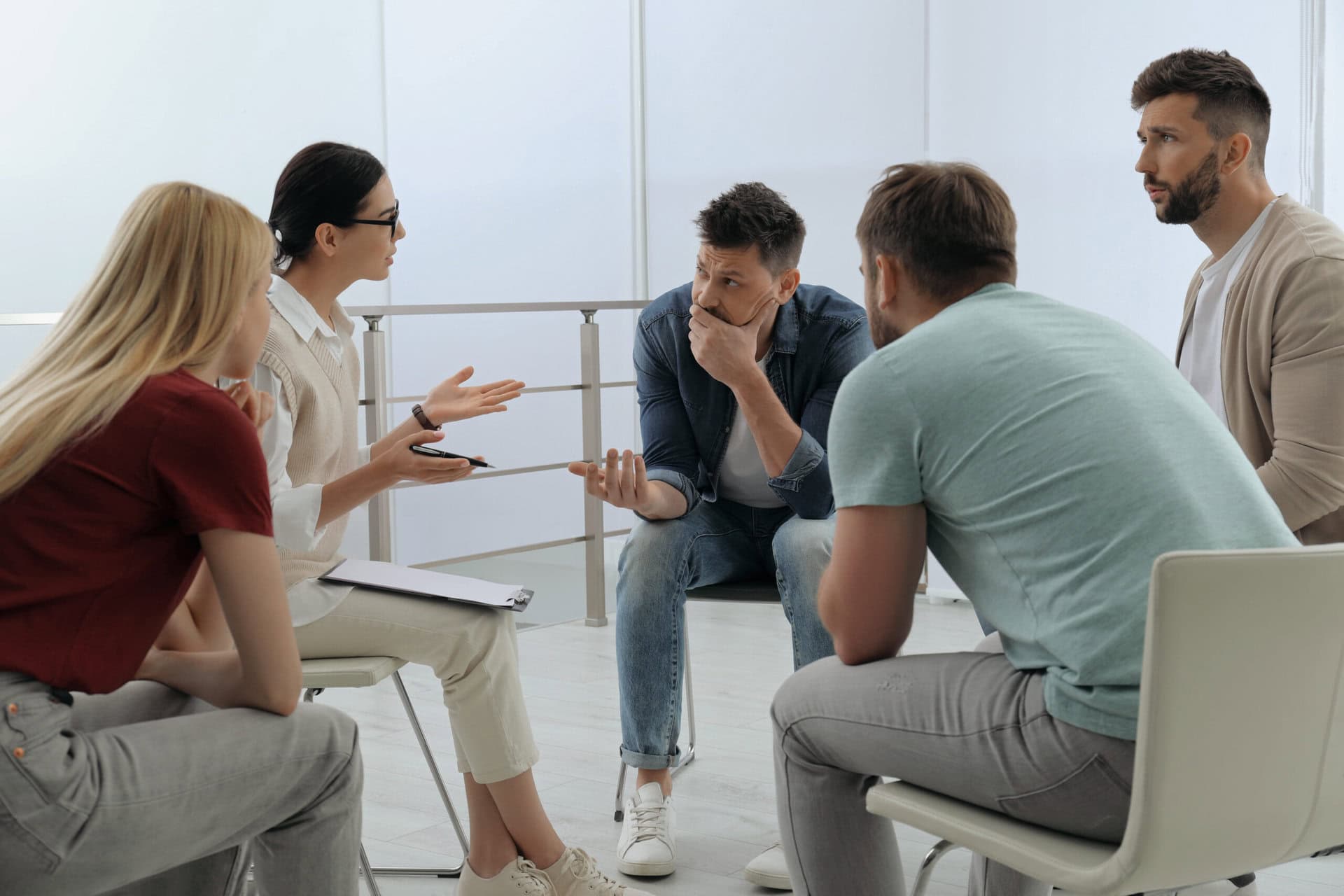 A group of five people sitting on chairs in a circle, engaged in discussion. A woman gestures with her hands while others listen attentively. The setting is a bright, modern room with light-colored walls and flooring.