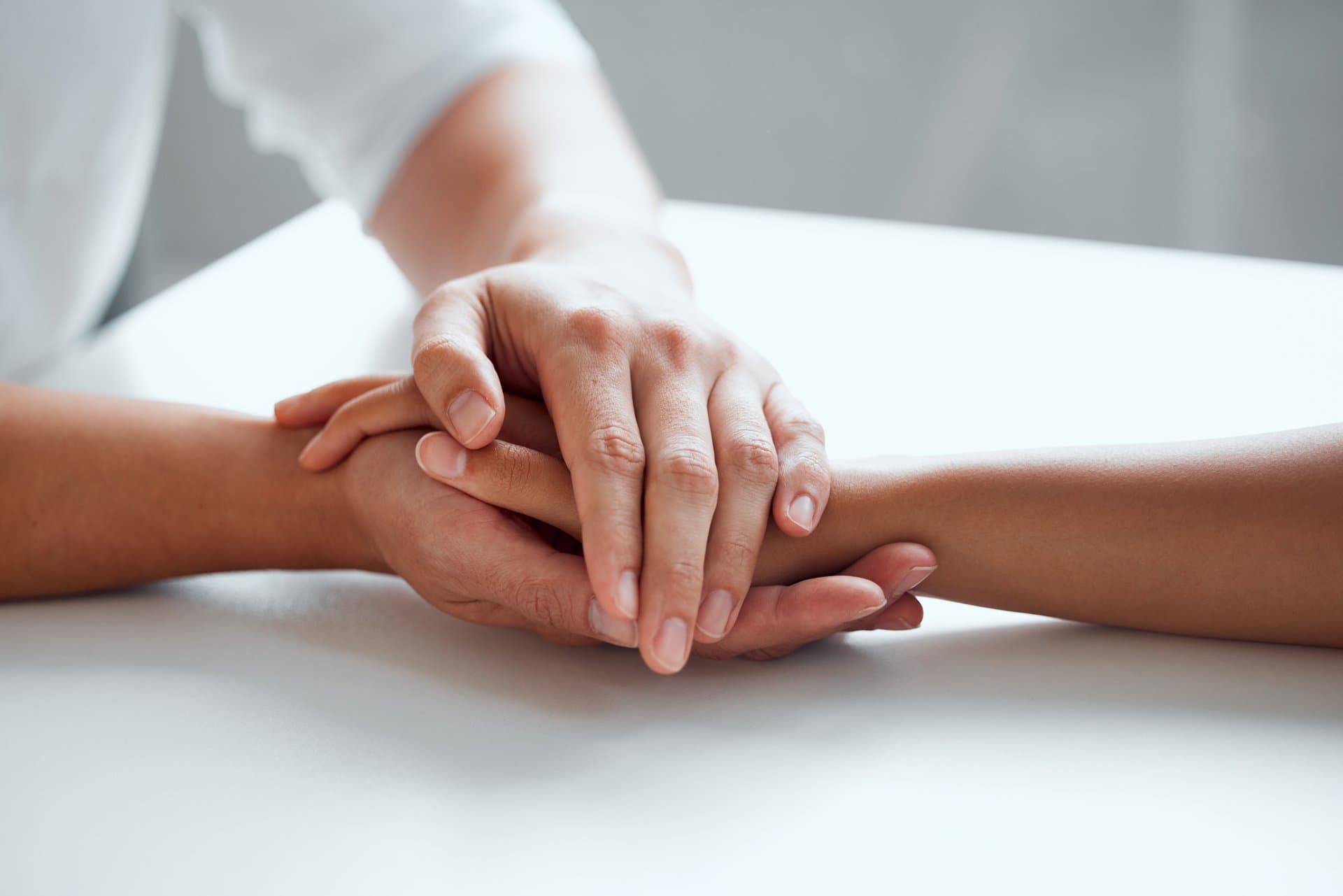 Two hands gently holding one another on a white table, symbolizing comfort and support. One persons hand is placed on top of the others in a comforting gesture.