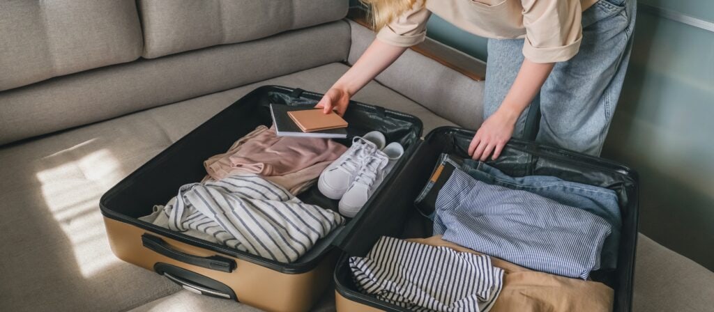 Person packing an open suitcase on a couch, with neatly folded clothes, sneakers, and holding a passport. The suitcase is half-packed, and light filters into the room, highlighting the casual, organized setting.