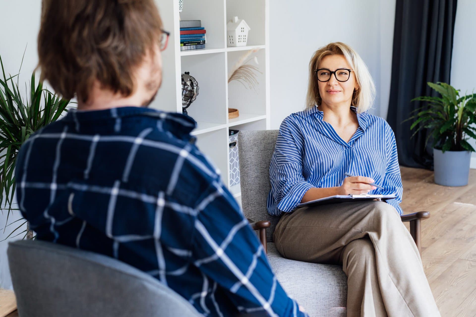 A woman in glasses and a blue striped shirt sits in a chair, holding a clipboard and pen, facing a man in a plaid shirt. They are in a bright room with a bookshelf and plants.