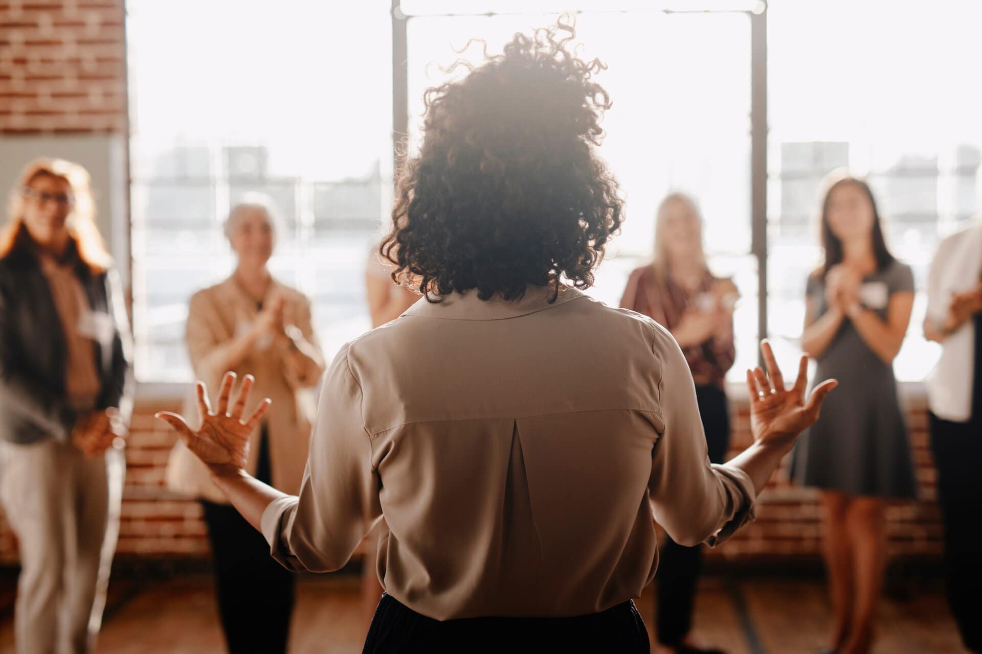 A person with curly hair stands facing a group in a sunlit room, gesturing with both hands. The group appears attentive and engaged, with some clapping. The room has large windows and exposed brick walls, suggesting an informal setting.