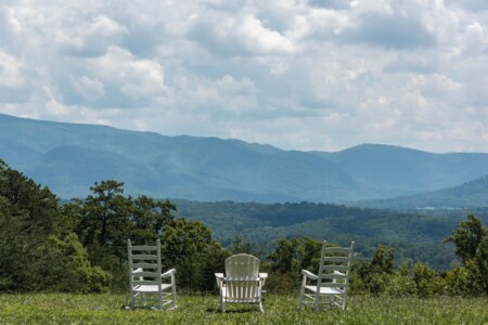 Three white chairs sit on a grassy hill overlooking a vast view of lush green mountains under a partly cloudy sky. The scene is tranquil and inviting, inviting contemplation of the natural beauty.