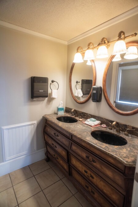 A bathroom with two oval mirrors, two sinks on a marble countertop, and a wooden vanity. Four pendant lights illuminate the space. A paper towel dispenser and soap dispenser are mounted on the beige wall. Beige tiled floor adds warmth.