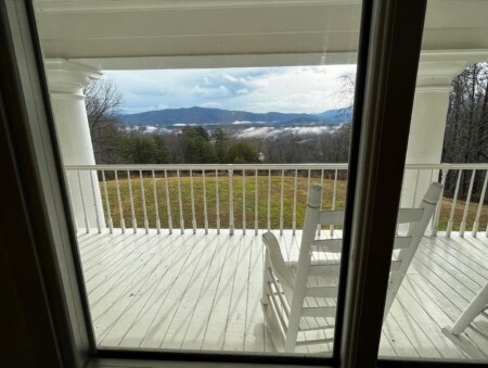View through a window of a white wooden porch with a rocking chair, overlooking a grassy field and distant mountains under a partly cloudy sky.