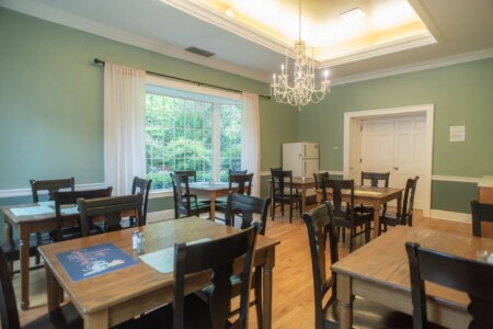 A dining room with green walls, a chandelier, wooden tables, and black chairs. Large window with white curtains lets in natural light. Wooden flooring and closed white double doors are visible in the background. Tables are set for dining.