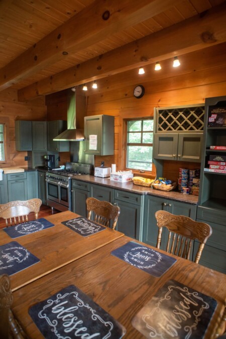 A cozy kitchen with wooden interiors features teal cabinets, a large dining table with blessed placemats, modern appliances, and counter space with cereal boxes and snacks. The room is warmly lit with natural and overhead lights.