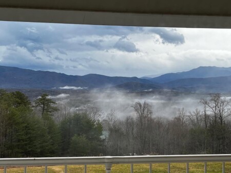 A scenic view of distant mountains under a cloudy sky, with mist rising from a forested area in the foreground. The scene is framed by the railing of a porch or balcony.