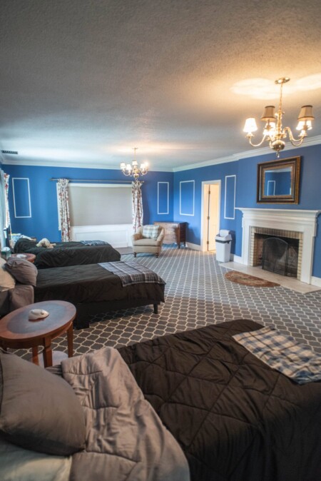 A cozy room with blue walls, featuring two beds with black bedding, a fireplace with a mirror above, a patterned carpet, and a cushioned chair by the window. Warm lighting from chandeliers creates a homey atmosphere.