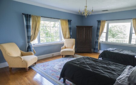 A cozy bedroom with blue walls features two twin beds with black bedding, two yellow armchairs, a dark wooden wardrobe, and large windows with blue plaid and yellow curtains. The room is bright with natural light coming in.