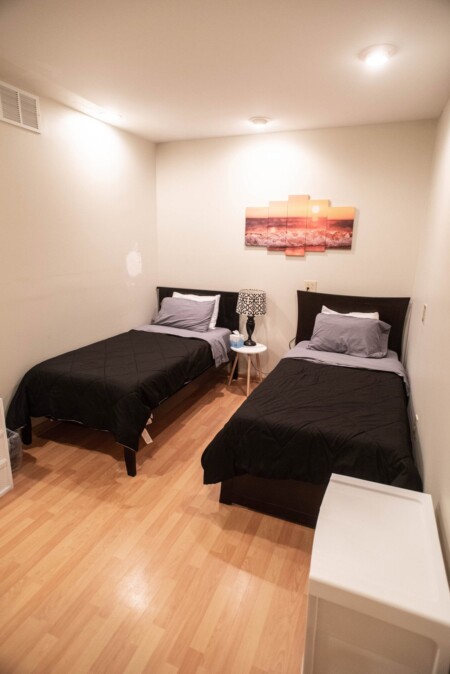 A minimalist bedroom with two single beds featuring black comforters and gray pillows. A small table with a patterned lamp sits between the beds. A multi-panel sunset artwork hangs on the wall above the beds. The floor is light wood.