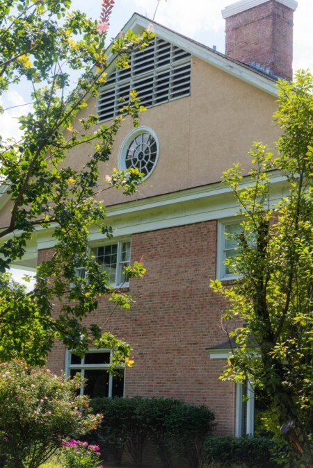 The image shows a two-story brick house with a circular window and a large chimney. Tall trees and lush greenery frame the house, partially obscuring its view. The sky is clear and blue.