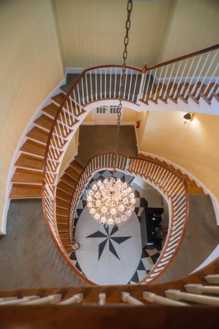 A spiral staircase made of wood with white railings curves around a sparkling chandelier. The chandelier hangs over a geometric tiled floor in a spacious, warmly lit interior.